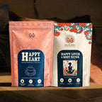 Two stand-up pouches of Sage & Soul herbal wellness products displayed side by side on a wooden surface in a dimly lit setting. The left pouch is peach-pink with a dark blue label reading 'Happy Heart Herbal Wellness Solution.
