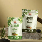 Two stand-up pouches of SAGE & NOLA plant-based nutrition products, 'Moringa Superfood' and 'Jasmine uplift', displayed on a stone surface with decorative plants and stones against a beige background.