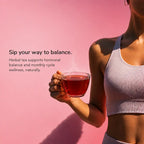 A woman in a ribbed light lavender sports bra and matching bottoms holds a clear glass mug filled with red herbal tea against a solid pink 