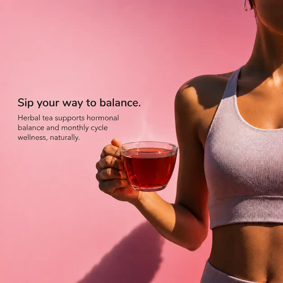 A woman in a ribbed light lavender sports bra and matching bottoms holds a clear glass mug filled with red herbal tea against a solid pink 