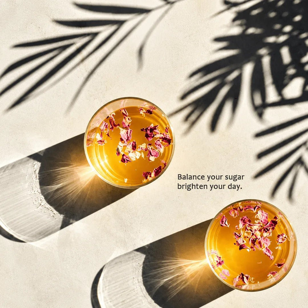 Two clear glasses filled with golden amber liquid and dried pink flower petals, arranged on a light beige surface with strong sunlight and palm leaf shadows. Black text in the center reads, 'Balance your sugar brighten your day.